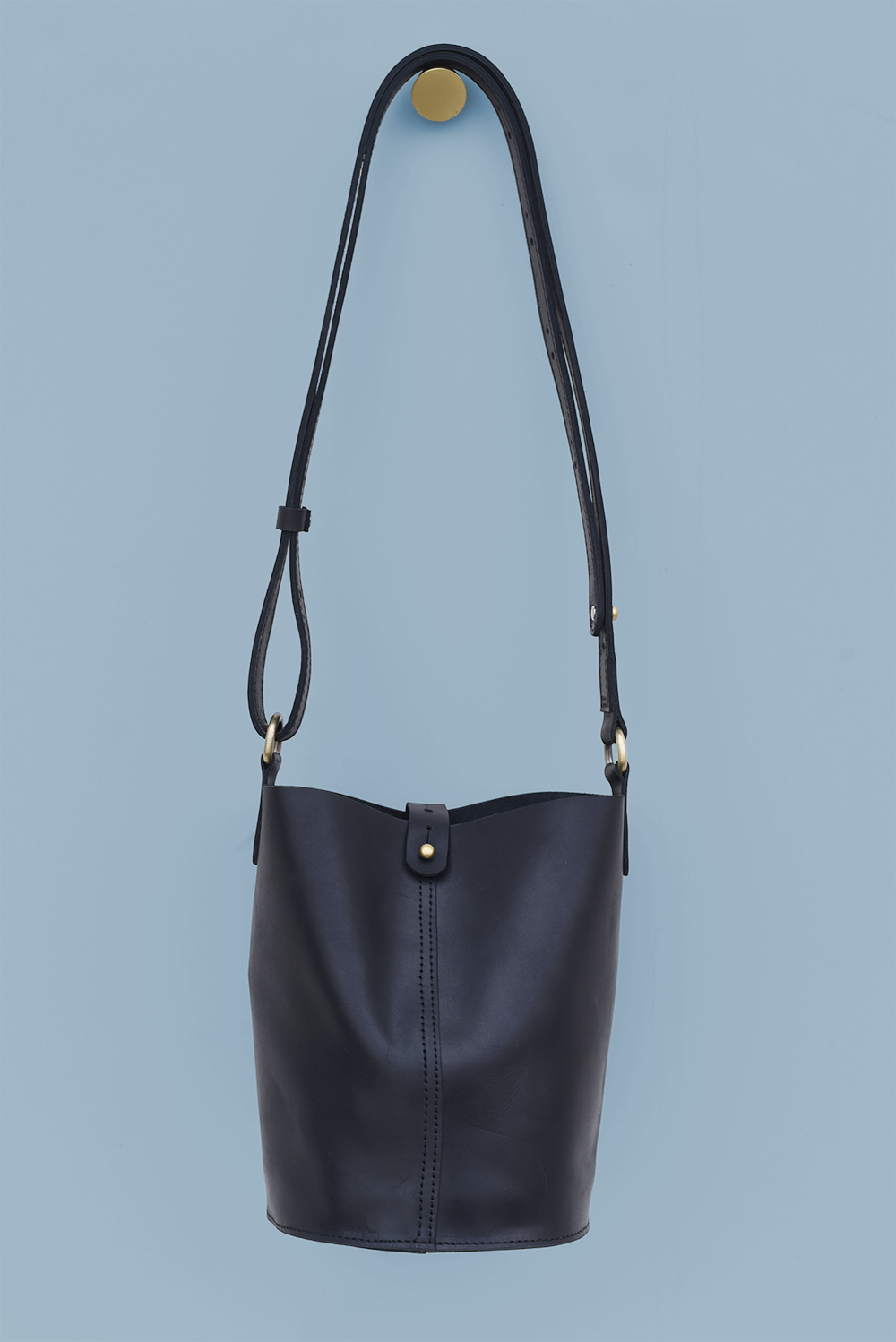 A black leather bucket bag with an adjustable crossbody strap and antique brass knob closure, displayed against a blue background.