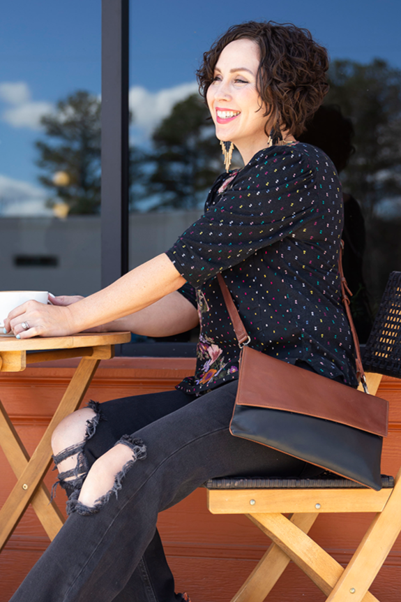 A person sitting outdoors wearing a black shirt with a pattern, blue jeans, and brown shoes, with a convertible crossbody and clutch bag on their lap.