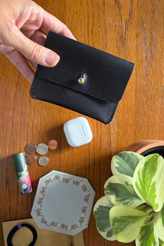 Black wallet held by a hand on a wooden surface with coins, airpods, and a plant.