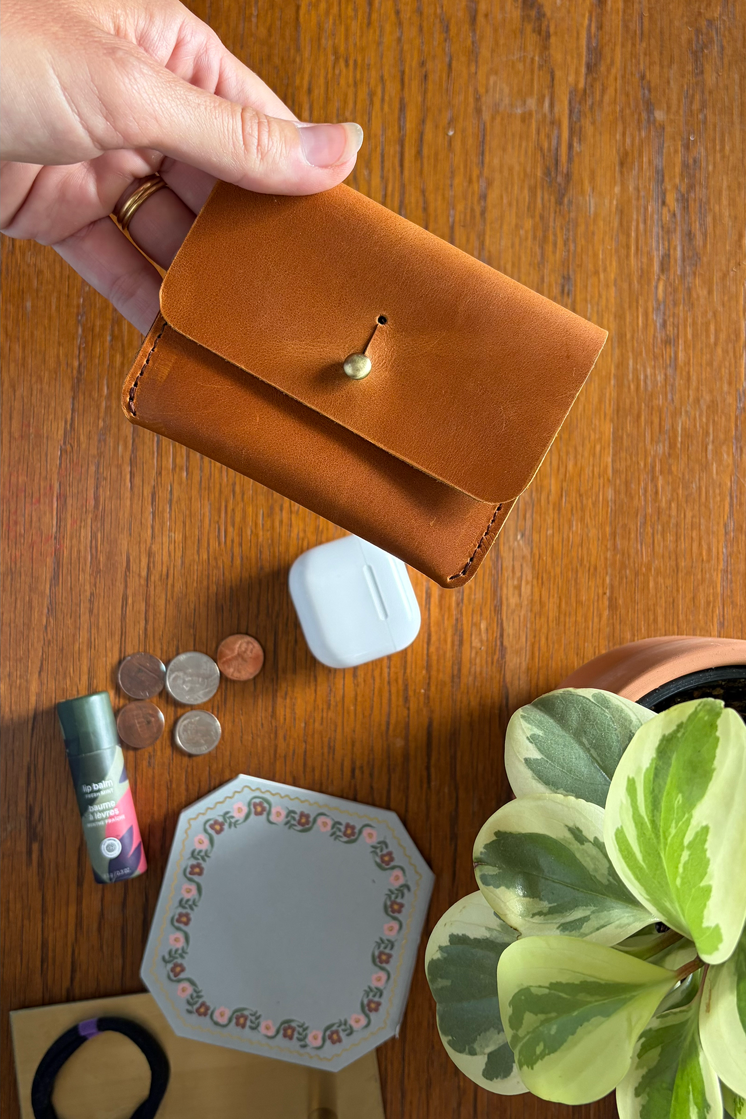 Brown leather card carrier held by a hand on a wooden surface with coins, airpods, and a plant.