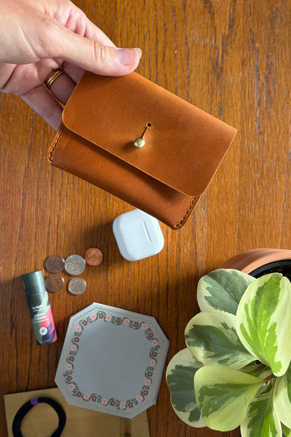 Brown leather card carrier held by a hand on a wooden surface with coins, airpods, and a plant.