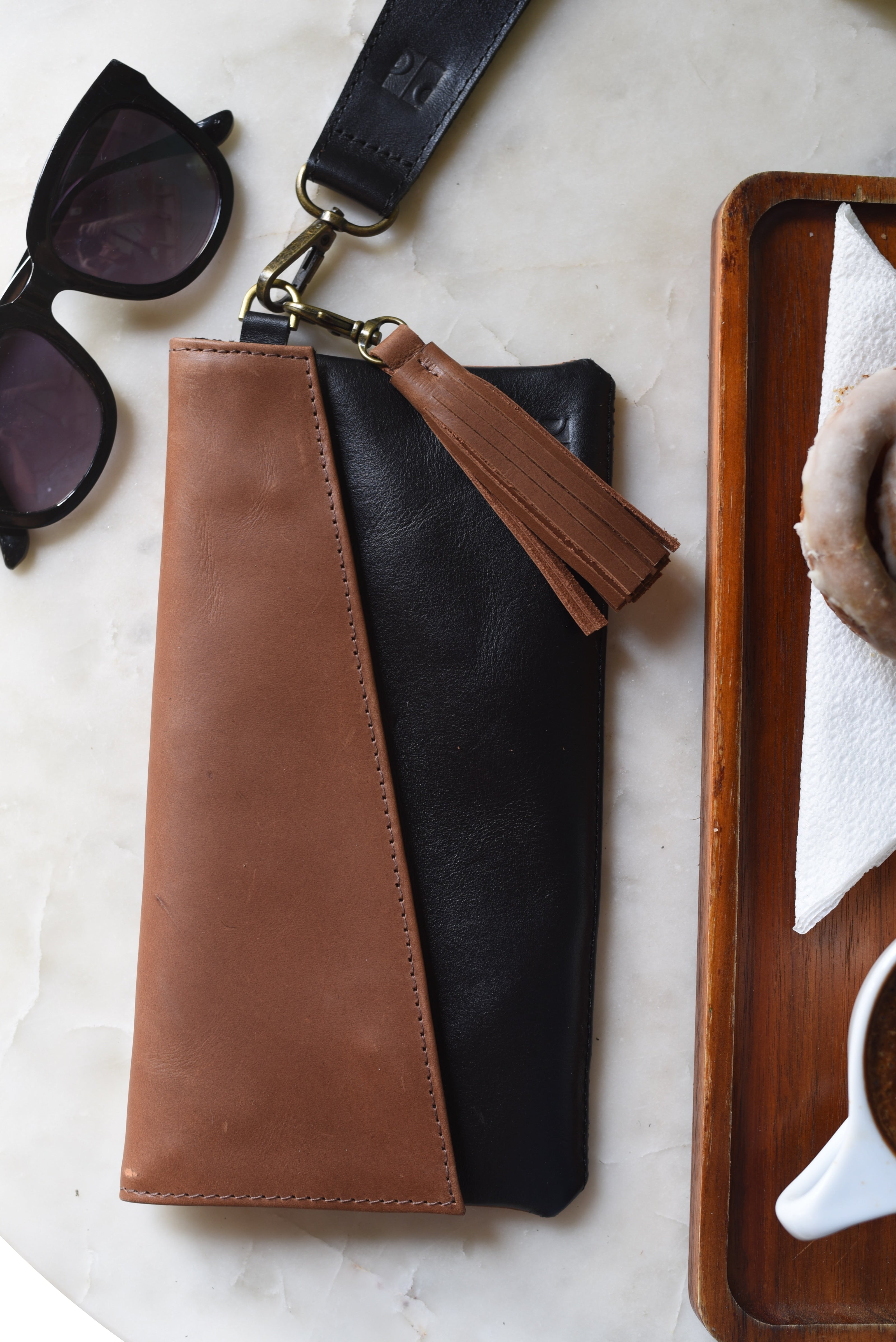 Brown and black leather clutch with tassel on a marble surface with sunglasses and a wooden tray.