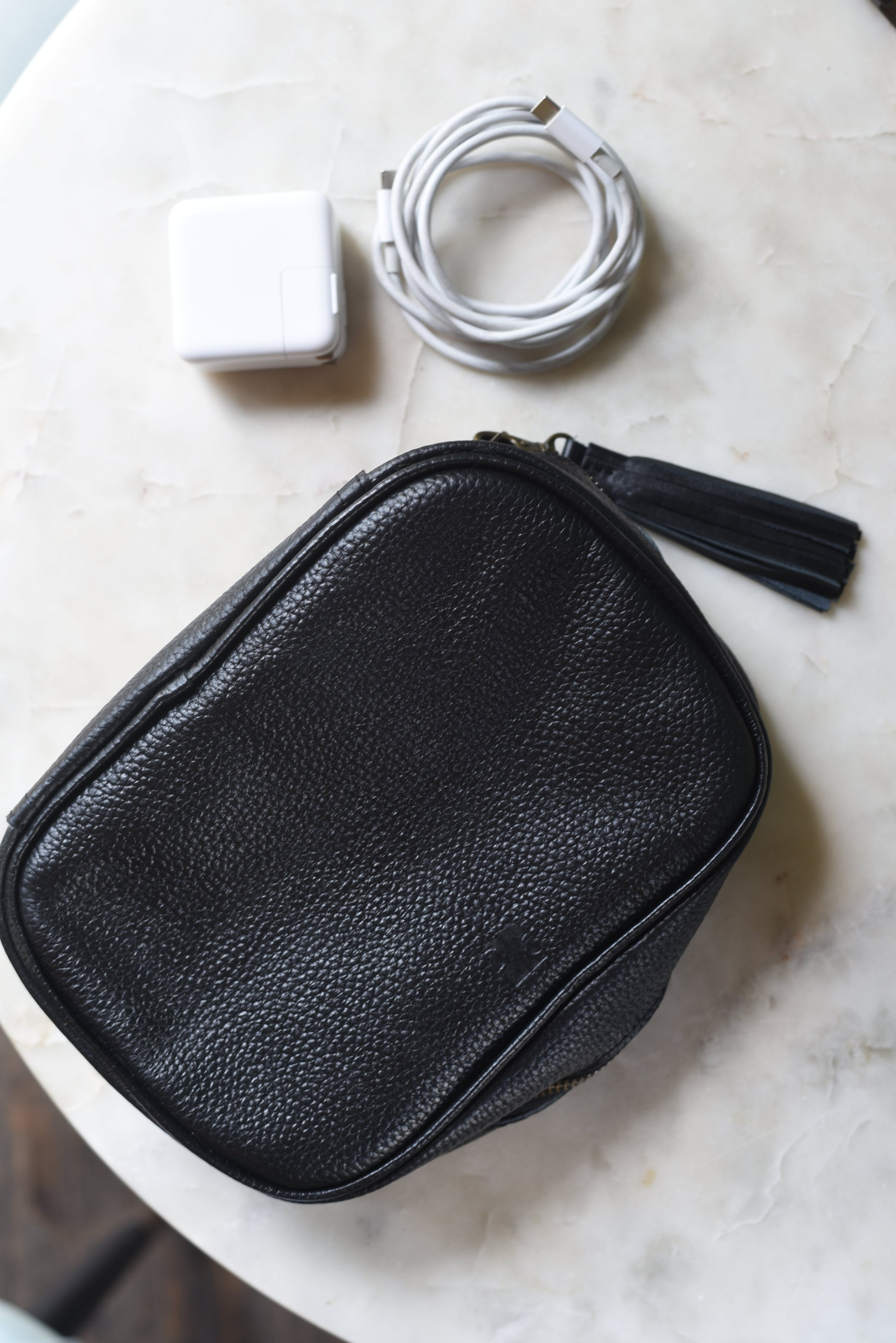 Black leather pouch with tassel on a marble surface, next to white power adapter and cable.