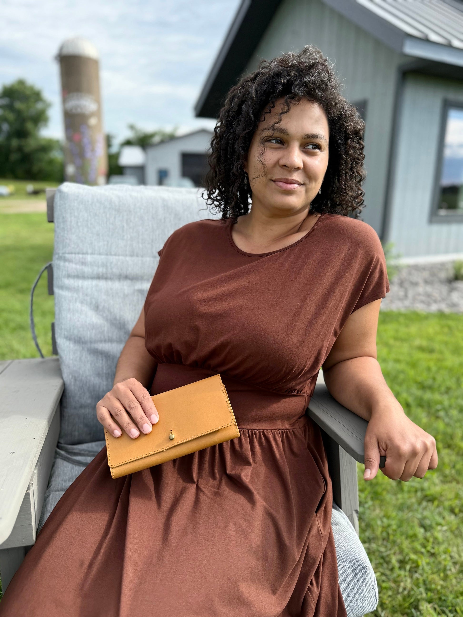 Woman in a brown dress holding a caramel leather wallet on a porch with a barn in the background