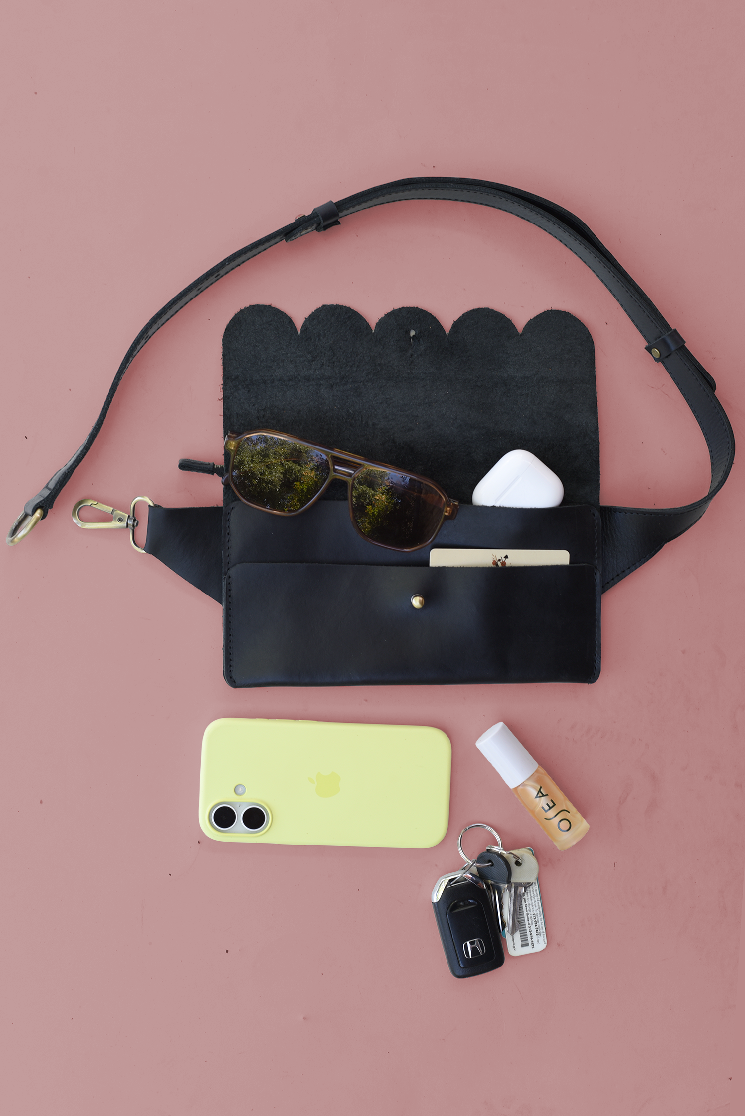 Black scalloped-edge bag with sunglasses, phone, keys, and lip balm on a pink background